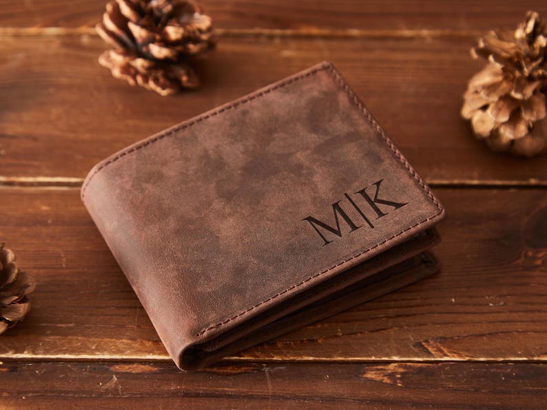 May include: A brown leather wallet with the initials "MK" embossed on the front. The wallet is rectangular and has a stitched border. The background includes pine cones on a wooden surface.
