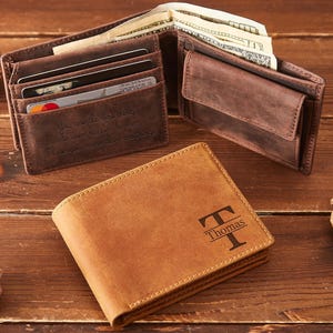May include: A brown leather wallet with the name "Thomas" and a stylized "T" engraved on the front. The wallet is open, revealing cash and card slots. Another open wallet and pine cones are also visible.