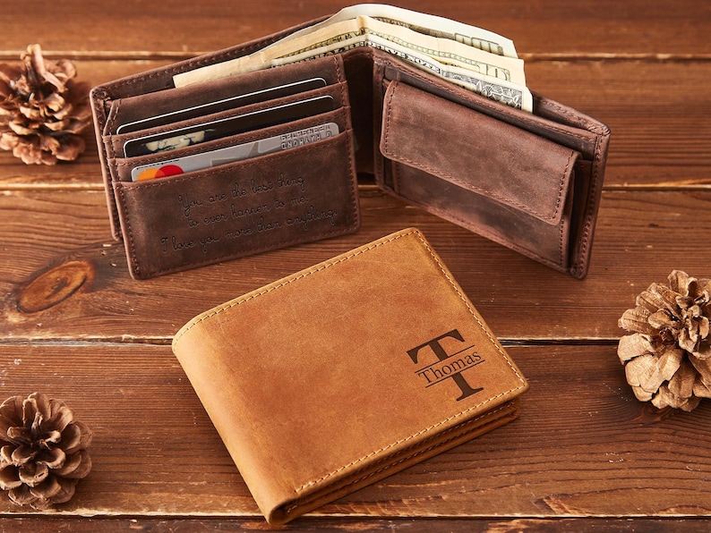 May include: A brown leather wallet with the name "Thomas" and a stylized "T" engraved on the front. The wallet is open, revealing cash and card slots. Another open wallet and pine cones are also visible.