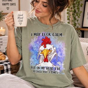 May include: A sage green t-shirt featuring a graphic of an angry white chicken with a red comb and yellow beak. The text reads, "I MAY LOOK CALM... BUT IN MY HEAD I'VE PECKED YOU 3 TIMES..." set against a purple and blue watercolor-style background.