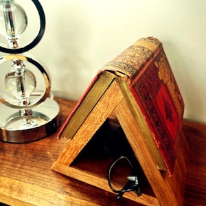 May include: A wooden triangular book stand with a book resting on top. The book is an older, hardback book with a red cover and gold lettering. The stand has a small shelf inside for holding glasses or other small items.