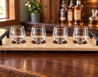 May include: A rectangular light-colored wooden serving tray featuring four circular cutouts designed to hold four clear glass snifters. The tray is equipped with two matte black metal handles on the ends and rests on small black feet.