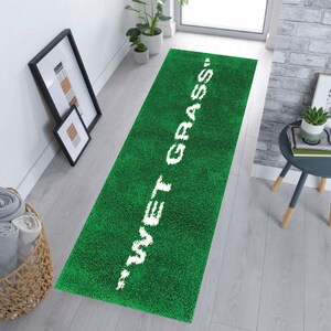 May include: A green rectangular rug with the words "WET GRASS" in white. The rug has a textured, grass-like appearance. The rug is placed in a hallway.