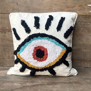 May include: Cream-colored square throw pillow featuring a large, embroidered eye design. The eye has a white center, a red iris, and is outlined with black, blue, yellow, and orange details. Black tufted lines radiate from the top.