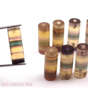 May include: A collection of eight multi-coloured gemstone cylinders, each with a different pattern of yellow, brown, green, and purple stripes. The cylinders are arranged in a row, with the largest cylinder on the left and the smallest cylinder on the right.