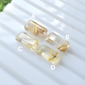 May include: Four rectangular, transparent quartz crystals with golden inclusions are arranged on a white surface. Each crystal is labeled with a letter, A, B, C, and D. The crystals vary in clarity and the density of the golden strands.