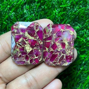 Può includere: Due pietre preziose rettangolari con un motivo a mosaico di magenta intenso e oro. Le pietre sono tenute in mano su uno sfondo di erba verde. Le pietre preziose hanno una superficie lucida e sfaccettata.