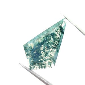 May include: A translucent gemstone, held by tweezers, with a distinctive pattern of green, tree-like inclusions. The stone's irregular shape is set against a white backdrop, accentuating its natural beauty. This gem is likely for jewellery making, with intricate details.
