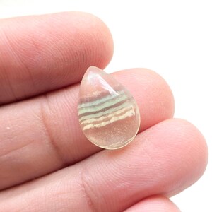 May include: A teardrop-shaped gemstone with layers of pink, green, and white. The polished stone is held in a hand, showing its translucent quality and natural patterns. The gemstone is likely used for jewellery or decorative purposes.