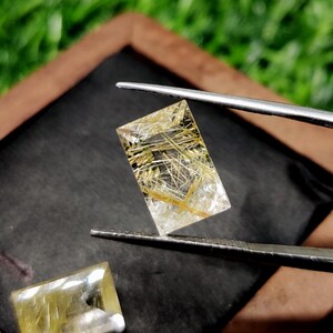 May include: A rectangular, transparent gemstone with golden, needle-like inclusions, held by silver tweezers. The gemstone is clear with a slight yellow tint, and the golden inclusions create a striking visual contrast. Another similar gemstone is visible in the lower left corner.