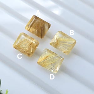 Pode incluir: Quatro pedras preciosas retangulares de quartzo rutilado dourado, rotuladas A, B, C e D. As pedras têm uma aparência translúcida com inclusões douradas em forma de agulha. As pedras são exibidas em uma superfície branca.