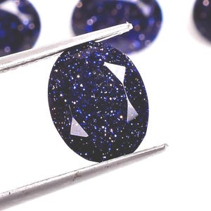 Peut inclure: Gros plan sur une pierre précieuse bleu foncé avec des inclusions scintillantes, tenue par une pince argentée. La pierre de forme ovale présente deux facettes triangulaires. D'autres pierres similaires sont visibles en arrière-plan, le tout sur fond blanc.