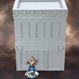 Könnte beinhalten: Ein zweistöckiges, hellgraues Gebäudemodell mit drei Fenstern im zweiten Stock und einem Schaufenster im ersten Stock. Eine Miniaturfigur eines Charakters mit Krallen und Hut steht vor dem Gebäude. Das Gebäude hat dekorative kreisförmige Akzente.