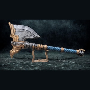 Op de afbeelding: Een gedetailleerde fantasy-bijl met een zilveren blad en gouden accenten. De handgreep is gewikkeld in blauw, ruitvormig materiaal. De bijl rust op een bijpassende standaard, die het ingewikkelde ontwerp en vakmanschap laat zien.