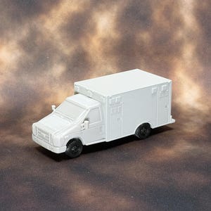 Può includere: Un piccolo modellino di ambulanza bianco non verniciato, caratterizzato da un vano posteriore squadrato, una cabina anteriore dettagliata con parabrezza, specchietti laterali e ruote nere. Il veicolo presenta dettagli esterni stampati e sagomati.