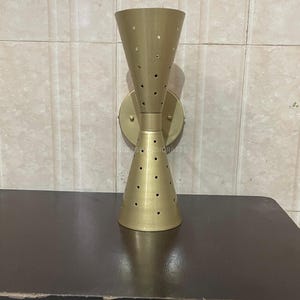 Puede incluir: Una lámpara de pared de color dorado con una forma de reloj de arena única y un diseño perforado. La lámpara tiene una base redonda y dos pantallas en forma de cono.