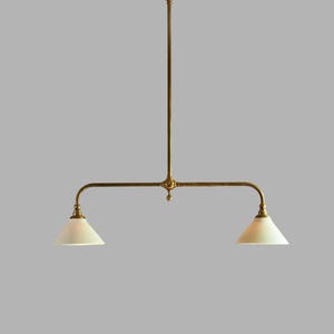 May include: A brass pendant light fixture featuring a central vertical rod connected to a horizontal bar with two downward-facing arms. Each arm supports a conical, off-white glass shade. The metal components have a warm, metallic finish.