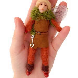 May include: A handmade fairy doll with a painted face, wearing an orange felt outfit with a green collar and a brown belt. The doll has a silver arrow charm and translucent wings. The fairy is approximately 10 cm tall.