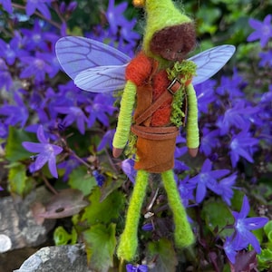May include: A needle-felted fairy ornament with a green pointed hat, brown hair, and translucent purple wings. The figure wears a brown leather-like vest and skirt over lime green felted arms and legs, suspended by a gold string.