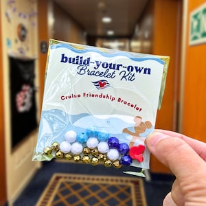 May include: A clear plastic package labeled "build-your-own Bracelet Kit" with the text "Cruise Friendship Bracelet." The kit contains various beads in white, blue, gold, and purple, along with red heart-shaped charms. The kit is held by a person.