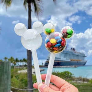 May include: Two clear plastic containers shaped like mouse heads with ears, each attached to a handle. One container is empty, while the other is filled with multicolored chocolate candies in shades of red, yellow, blue, green, and brown.