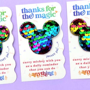 May include: A mouse-shaped patch covered in iridescent, multi-colored sequins attached to a white card. The card features the text "thanks for the magic" and "carry mickey with you as a daily reminder that you can do anything" with a "pocket pal" label.