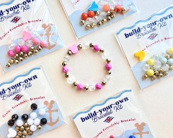 May include: A collection of build-your-own bracelet kits in clear plastic packaging, each containing colorful beads, metallic gold beads, and a mouse-shaped bead. A finished bracelet with pink, clear, and gold beads sits in the center.