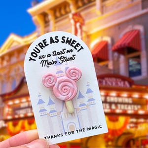 May include: A pink Mickey Mouse-shaped lollipop on a white card with the text "YOU'RE AS SWEET as a treat on Main Street" and "THANKS FOR THE MAGIC". The lollipop has a swirl design and is on a light-colored stick.