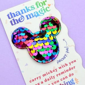 A close-up view of a rainbow sequin Mickey mouse shape attached to a white card that reads thanks for the magic.
