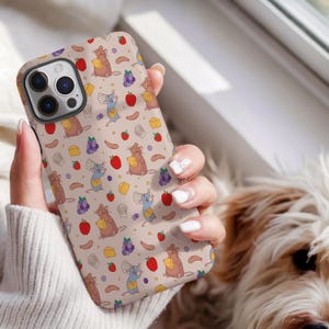 Puede incluir: Funda de teléfono beige con un patrón repetido de ratones de dibujos animados, queso, uvas, manzanas y salchichas. La funda tiene un diseño protector con un recorte para la cámara y está sujeta por una persona.