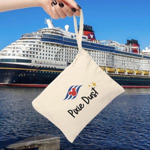 May include: A cream-colored canvas pouch with a handle, featuring the words "Pixie Dust" and a logo in blue, red, and yellow. The pouch is held up in front of a large cruise ship on a sunny day.
