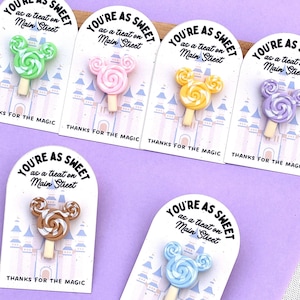May include: Six lollipop-shaped pins in green, pink, yellow, purple, brown, and blue, each featuring a Mickey Mouse-inspired swirl design. Each pin is attached to a white card printed with a castle graphic and the text, You're as sweet as a treat on Main Street, thanks for the magic.