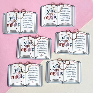 May include: Bookmarks with a book design, featuring the words "You are magic" in burgundy and blue. Each bookmark has a bow-shaped clip and includes the text "Thanks for being a part of our story. Wishing you a year filled with magic."