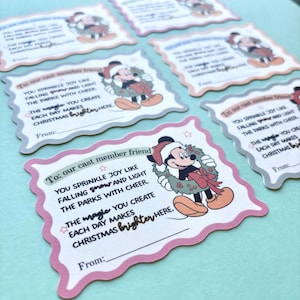 May include: Several Christmas cards with a pink and gray scalloped border are arranged on a light blue surface. Each card features Mickey Mouse wearing a Santa hat and holding a wreath. Text on the cards reads, "To our cast member friend" and a Christmas message.