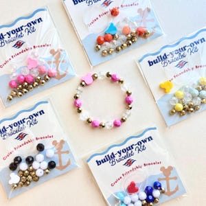 May include: A completed bracelet with pink, clear, and gold beads sits among several "build-your-own bracelet kit" packages. The kits contain various colored beads, charms, and elastic string. The text on the packages reads "Cruise Friendship Bracelet".
