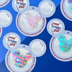 May include: Close-up of several cruise-themed gift tags with a Mickey Mouse head design made of colorful sequins. The tags have the text "Your Cruise Par" and "Here to remind you that you can do anything! happy sailing, friends!" on a blue background.
