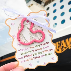 May include: A pink Mickey Mouse-shaped keychain attached to a decorative tag with a white ribbon. The tag has a scalloped edge and features a world map and the text "wherever you may wander...may Mickey journey with you and safely bring you home."