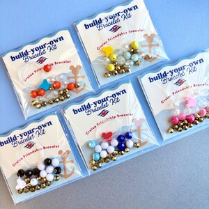 Five DIY bracelet kits in individual plastic packaging are displayed against a light blue surface, showing varied bead colors.