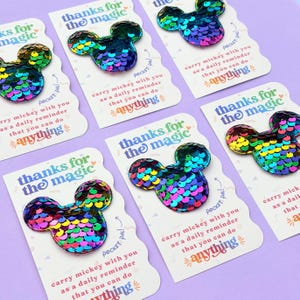 Several white cards with scalloped edges and rainbow sequin Mickey mouse shapes are scattered on a purple surface.