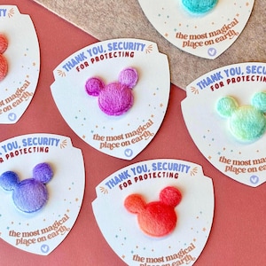May include: A collection of colorful Mickey Mouse-shaped patches on white shield-shaped cards. Each patch is a different color, including pink, blue, and orange. The cards read "THANK YOU, SECURITY FOR PROTECTING" and "the most magical place on earth."