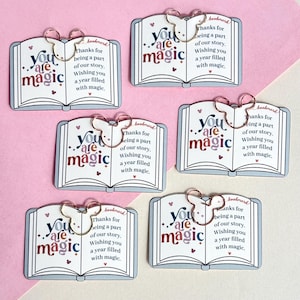 May include: Six rectangular paper bookmarks shaped like open books, each featuring a rose gold-colored metal paperclip in the shape of mouse ears. The text reads: you are magic, thanks for being a part of our story, wishing you a year filled with magic.