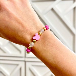 A pink beaded bracelet with gold spacers and a pink Mickey bead is worn on a person's wrist.