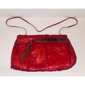 May include: A bright red handbag featuring a gathered, pleated body and a thin shoulder strap. The bag is accented with a horizontal band of dark red, snakeskin-textured material that ties into a decorative bow on the left side of the front.