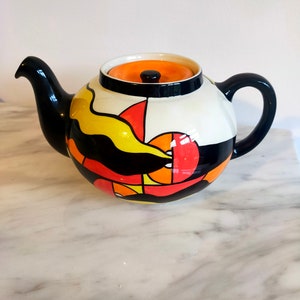 Könnte beinhalten: Eine schwarz-weiße Teekanne mit einem geometrischen Muster in Orange und Gelb. Die Teekanne hat einen Deckel mit einer orangefarbenen Oberseite und einem schwarzen Knopf.