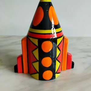 May include: A ceramic rocket-shaped salt and pepper shaker with a black, orange, and yellow geometric design. The shaker has a cone-shaped top and a rounded base.