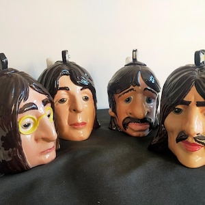 Può includere: Quattro sculture di teste in ceramica, ognuna raffigurante un membro di una famosa band. Le sculture sono dipinte con tonalità della pelle realistiche, capelli e lineamenti del viso. Una scultura ha occhiali con montatura gialla. Le teste sono disposte su una superficie scura.