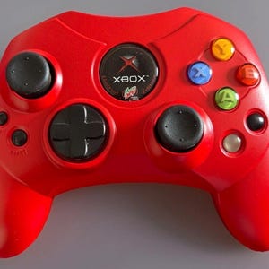 May include: A red Xbox controller with black joysticks, a directional pad, and colored action buttons. The controller features the Xbox logo and the text "Limited Edition".