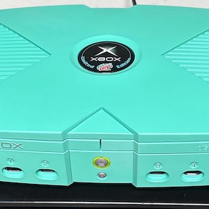 Puede incluir: Una consola Xbox de edición limitada de color turquesa. La consola tiene un botón de encendido circular y cuatro puertos para mandos. El logotipo de Xbox está centrado en la parte superior de la consola.