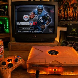 May include: A translucent orange Xbox video game console and matching controller with internal lighting, positioned next to a can of Mountain Dew LiveWire. A JVC television displays the Madden NFL 07 title screen, with Xbox game cases stacked nearby.