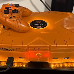 May include: Orange Xbox console and controller. The console features a transparent design, with the Xbox logo on top. The controller sits on the console. Both the console and controller are illuminated with orange lights.